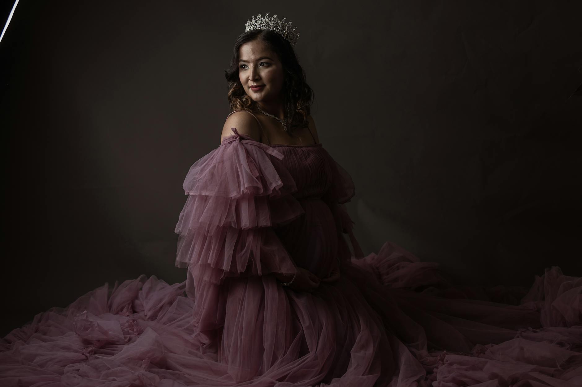Kobieta w tiulowej sukni i koronie pozuje w studyjnym, matowym świetle